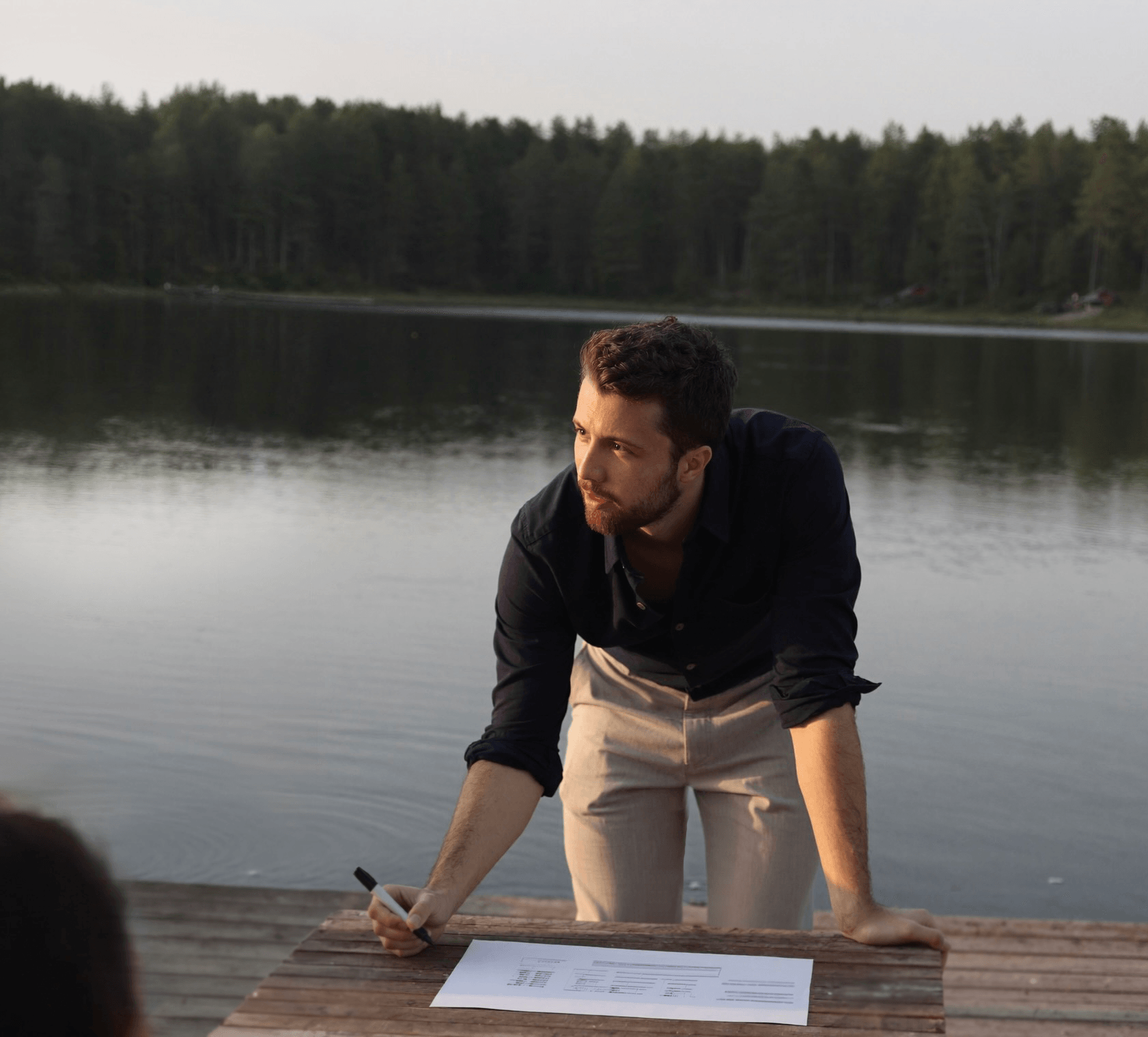 Julius leaning over a sheet of paper on a lakeside jetty, marker in hand, looking up from the page.