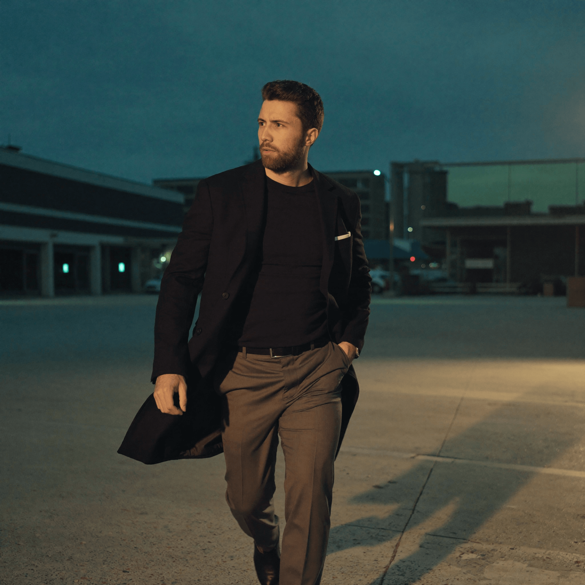 Julius walking across an empty parking lot at dusk in a long coat.